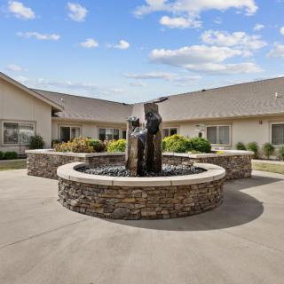 A courtyard with a central fountain surrounded by a stone border, set against a building under a blue sky with clouds.
