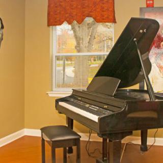 A grand piano sits in a cozy room with warm lighting, featuring a window and artwork on the wall.