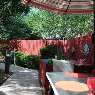 A charming outdoor patio area with a red umbrella, surrounded by lush greenery and a wooden fence.