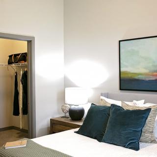 A modern bedroom featuring a neatly made bed with teal pillows, a bedside lamp, and abstract artwork on the wall. An open closet is visible to the side.