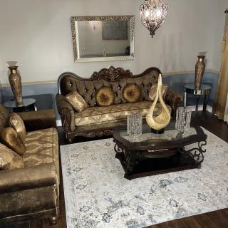 A traditionally decorated living room featuring an ornate sofa set, a central coffee table with decorative pieces, and elegant vases on either side. A large mirror hangs above the sofa, complemented by a chandelier.