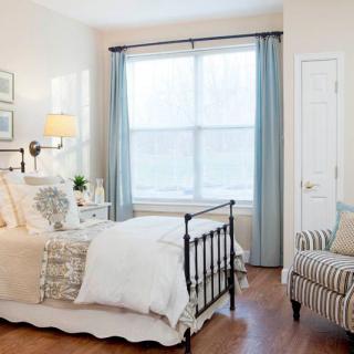A cozy and bright bedroom featuring a neatly made bed, soft lighting from a bedside lamp, and large windows with pale blue curtains.