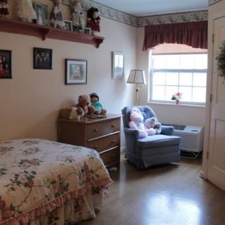A cozy bedroom featuring a bed with floral covers, shelves with dolls and toys, and a comfortable chair by the window.