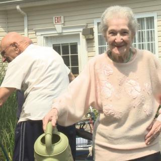 An elderly woman smiles while watering plants in a backyard garden.