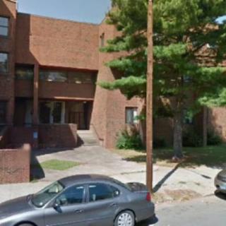 A brick building with parked cars and a tree along the sidewalk.