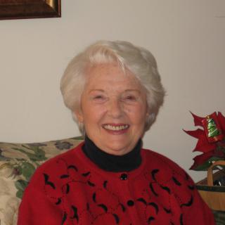 An elderly woman with white hair is smiling indoors, wearing a red sweater.