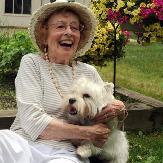 An elderly woman wearing a hat is joyfully holding her dog in a garden surrounded by flowers.