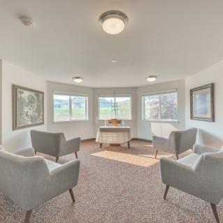 A cozy room with four gray chairs arranged around a central table, surrounded by artwork and windows offering views.