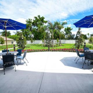 A sunny outdoor patio area with tables, chairs, and blue umbrellas overlooking a landscaped garden.