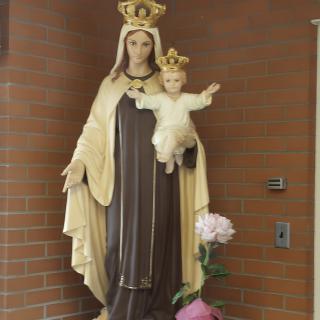 A statue of the Virgin Mary holding a child, adorned with crowns, set against a brick wall with a flower placed nearby.