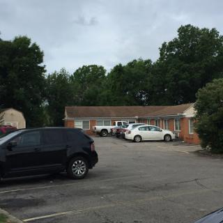 A view of a motel parking lot with several cars parked and surrounded by trees under a cloudy sky.