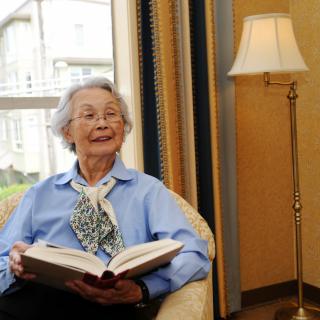 An elderly woman sitting comfortably in a chair, reading a book, with a lamp and window in the background.