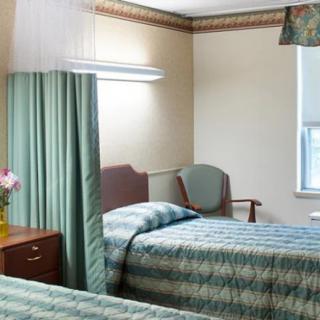 A hospital room with two beds separated by a curtain, featuring a chair and flowers by the window.