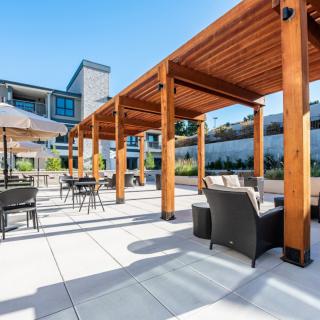A modern outdoor patio with a wooden pergola, comfortable seating, and tables under a clear blue sky.