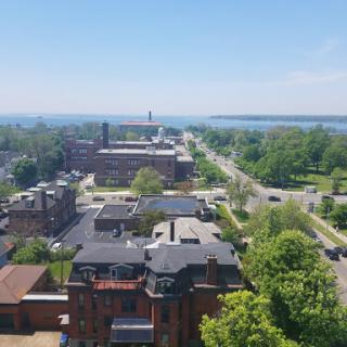 A panoramic view of a city overlooking a waterfront, featuring lush greenery and a clear blue sky.