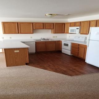 A spacious kitchen with wooden cabinets, white appliances, and a mix of carpet and wood flooring.