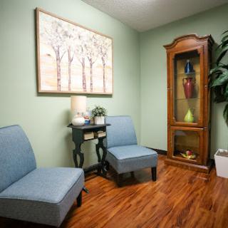 A cozy waiting room featuring two blue chairs, a small table, and a decorative cabinet against green walls.