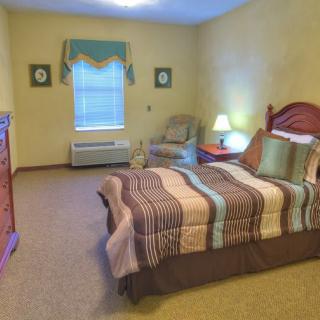 A cozy bedroom interior featuring a bed with colorful bedding, a bedside lamp, and an armchair by the window.