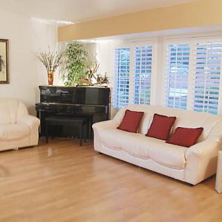 A cozy living room with a sofa, armchair, and piano, featuring hardwood floors and large windows with shutters.