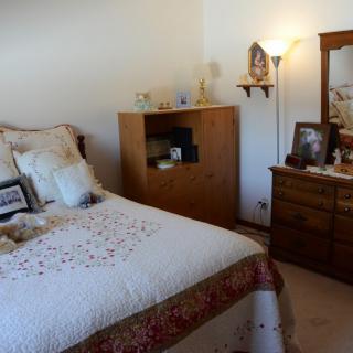 A cozy bedroom featuring a bed with decorative pillows, a wooden dresser with a mirror, and ambient lighting from a floor lamp.