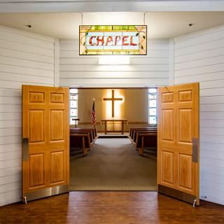 An inviting chapel with wooden doors open, revealing a cross at the front and rows of seating inside.