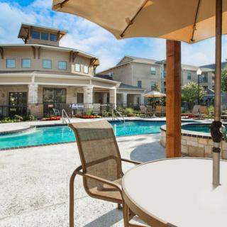 A sunny outdoor swimming pool area with patio furniture and umbrellas in a residential setting.