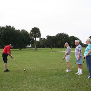 A group of seniors watches as a younger golfer demonstrates a swing technique on the green.