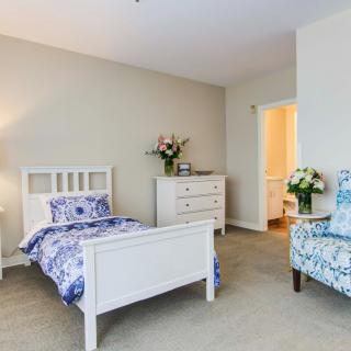 A cozy bedroom with a single bed, decorated with floral bedding, an armchair, and a dresser topped with fresh flowers.