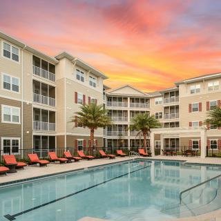 A modern apartment complex featuring a swimming pool surrounded by palm trees at sunset.