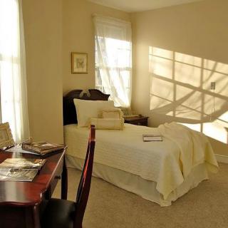 A cozy bedroom with a single bed and sunlight streaming through the window onto a desk.