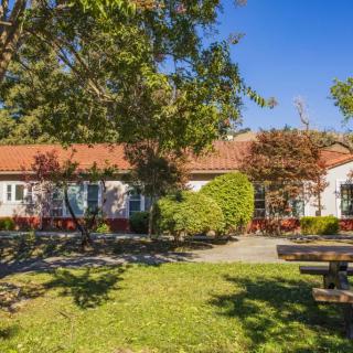 A serene outdoor scene featuring a building with a red-tiled roof, surrounded by trees and a well-maintained lawn under a clear blue sky.