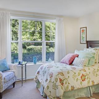 A cozy bedroom with a large window allowing natural light to fill the space, featuring a bed with floral-patterned bedding and an armchair.