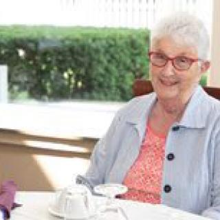 An elderly woman with glasses is smiling while sitting at a dining table indoors.
