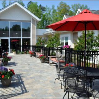 A charming outdoor patio area with tables and chairs, shaded by red umbrellas, surrounded by flowers and greenery.