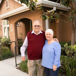 An elderly couple smiling and standing together in front of their house.