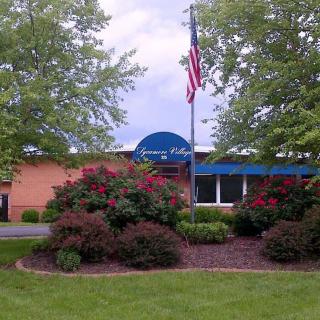 A building entrance with surrounding landscaping, featuring trees, a garden, and an American flag.