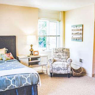 A cozy bedroom with a neatly made bed, an armchair, and a lamp by the window.