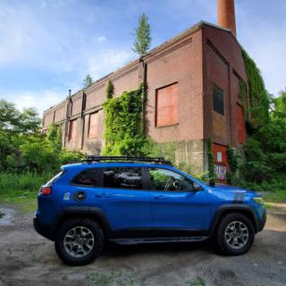 A blue SUV parked next to an overgrown, abandoned brick building under a clear sky.