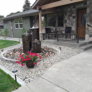 A well-maintained front yard featuring a rock garden with a fountain and vibrant flowers, adjacent to a stone house with outdoor seating.