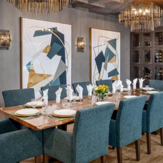 A modern dining room setting with a large table, blue chairs, abstract artwork, and elegant chandeliers.