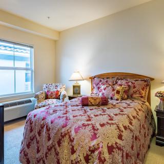 A cozy bedroom featuring a bed with floral bedding, a side table with lamps, and a window allowing natural light to brighten the space.