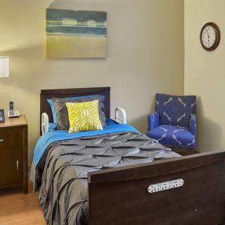 A cozy bedroom featuring a single bed with colorful pillows, a side table with a lamp, and a comfy chair next to it. A wall clock and painting decorate the space.