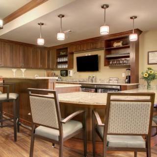 A modern kitchen interior with wooden cabinets, a central island with chairs, pendant lighting, and a wall-mounted television.