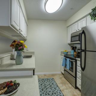 A modern kitchen with stainless steel appliances and white cabinets, featuring a decorative rug and flowers on the counter.