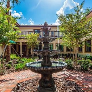 A tranquil courtyard featuring a central fountain surrounded by lush greenery and a two-story building under a bright blue sky.
