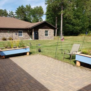 A sunny garden patio with raised flower beds and a view of a house surrounded by grass and trees.
