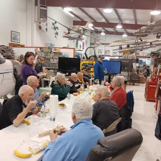 A community gathering of elderly individuals enjoying a meal together in an aviation-themed indoor space.