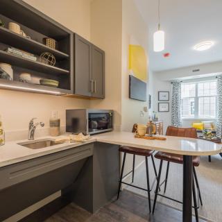 A modern kitchen and dining area in an apartment, featuring a countertop with stools, a microwave, and open shelving.