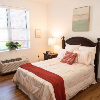 A cozy bedroom with a neatly made bed featuring red and white bedding, a bedside lamp, and a window bringing in natural light.