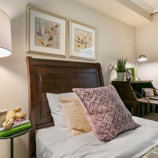 A cozy bedroom with a neatly made bed, decorative pillows, artwork on the wall, and a bedside lamp.